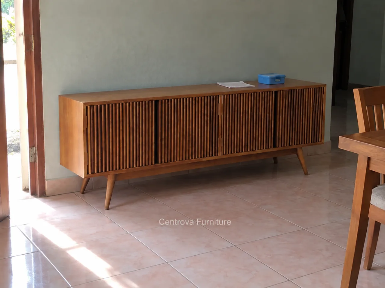Buffet jati berpintu bilah vertikal dalam ruang makan sederhana dengan cahaya dari pintu.